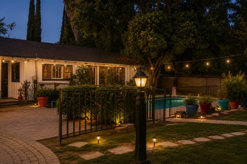 Nighttime backyard patio with illuminated house, metal fence, landscape lighting, and pool area surrounded by trees
