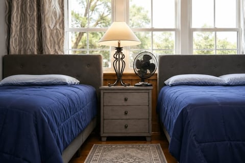 Twin bedroom with blue bedding, wooden nightstand with lamp and fan, large windows with curtains, and hardwood flooring.