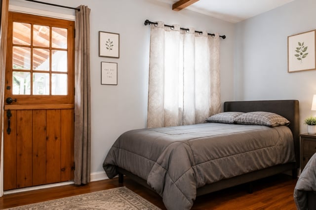 Bright bedroom with a dark bed, wooden door, white curtains, framed botanical prints, and wood flooring.