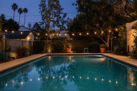 Illuminated backyard swimming pool at dusk with string lights, palm trees, and residential buildings in evening ambiance