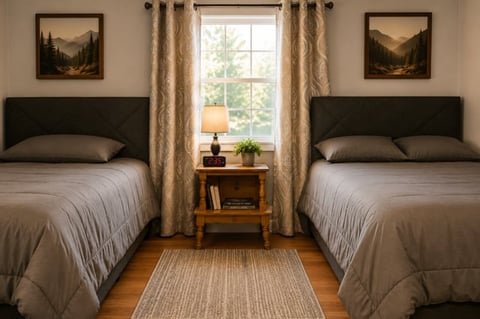 Symmetrical bedroom with two twin beds, dark headboards, beige bedding, wooden nightstand with lamp between beds, window with tan curtains, and mountain artwork on walls