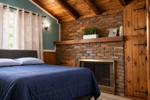 Cozy bedroom with blue-green walls, wooden ceiling, exposed brick fireplace, dark blue bedding, and wooden door with decorative hardware