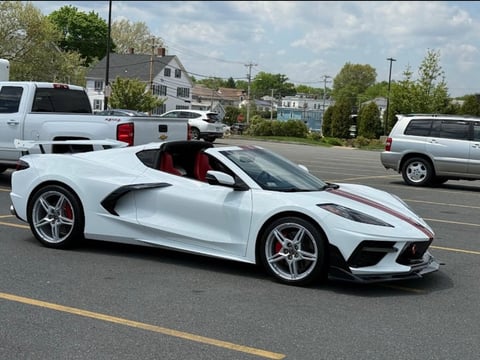 White sports car with red racing stripes parked in a lot with other vehicles and houses in background