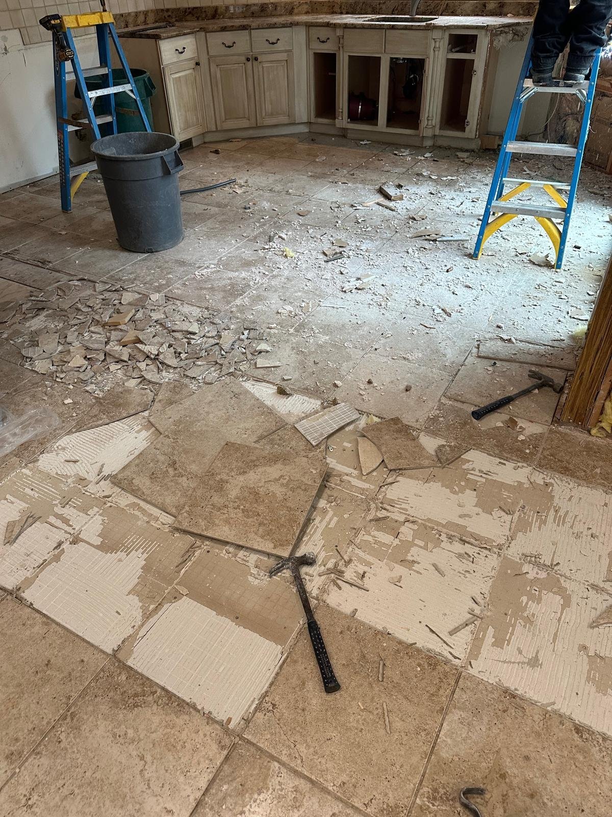 Kitchen renovation in progress with removed flooring, cream cabinets, blue ladders, and construction debris scattered on concrete subfloor