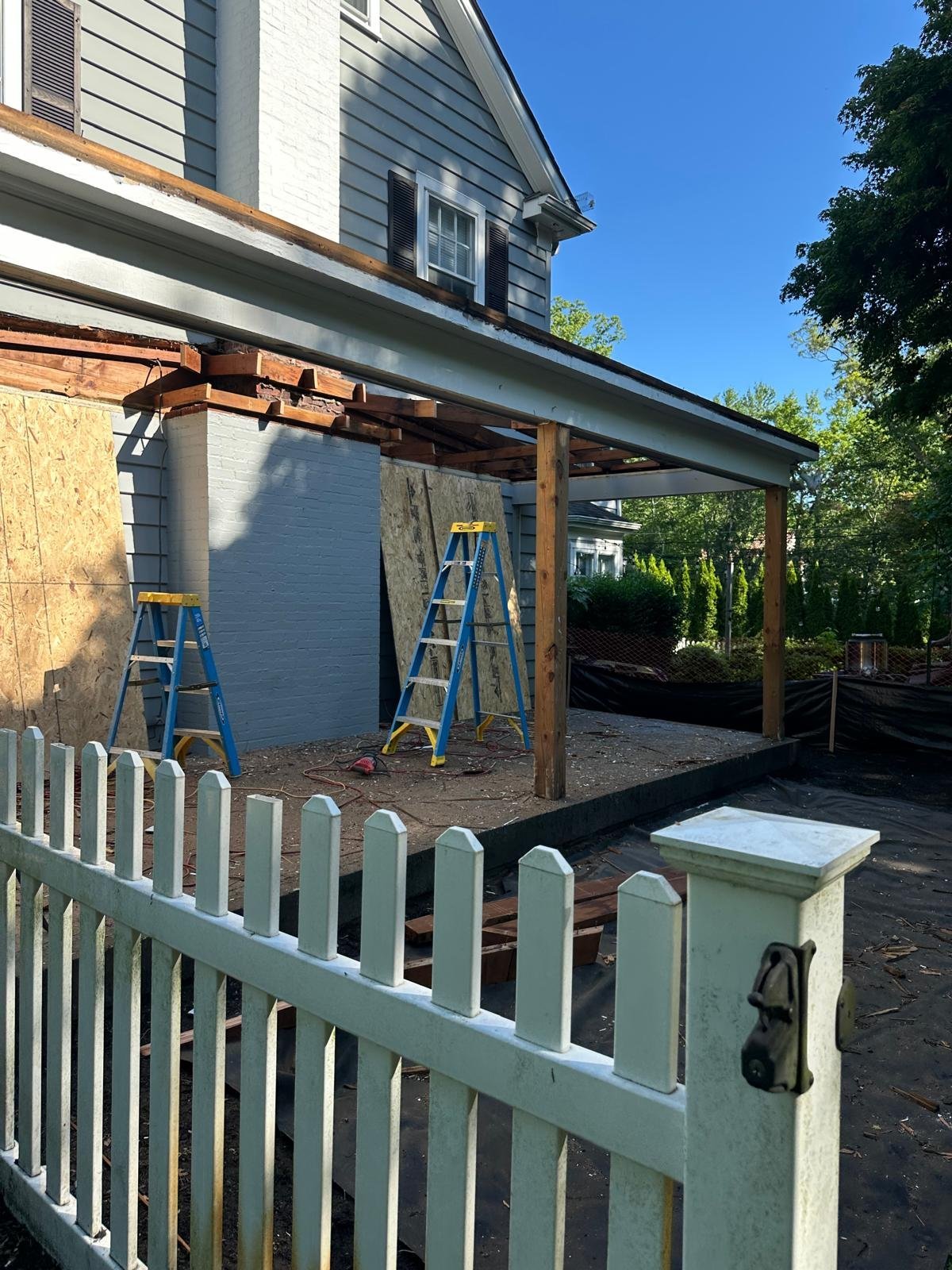 Home renovation with wooden pergola structure, blue ladders, white picket fence, and partially constructed addition to residential house under clear sky