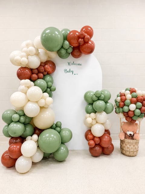 Baby shower backdrop with Welcome Baby sign, arch of white cream and sage green balloons with rust accents, and teddy bear decoration