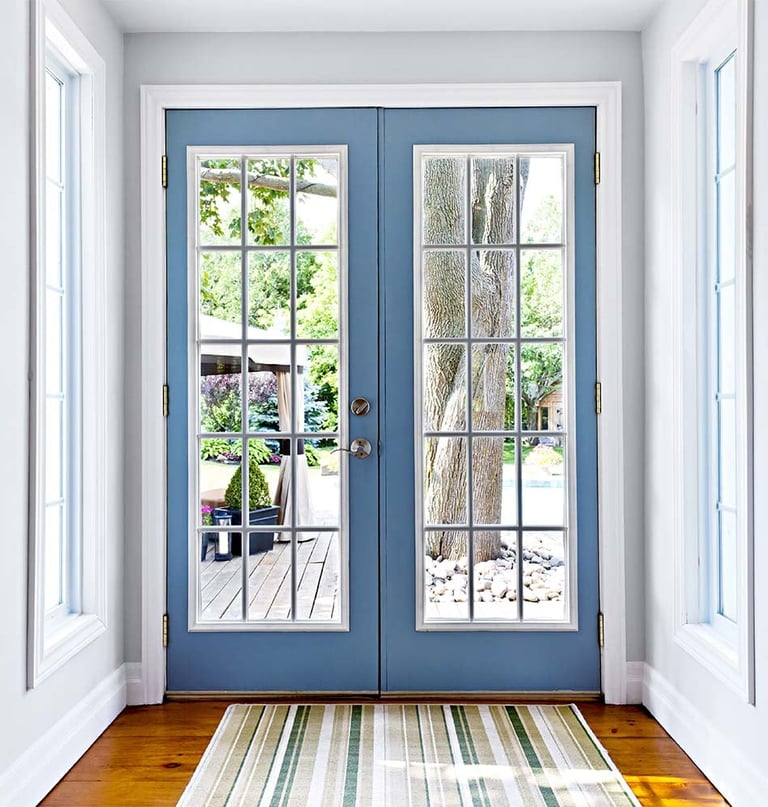 Pair of blue double doors with glass panes opening to a patio, flanked by white side windows in a bright hallway with hardwood floors.