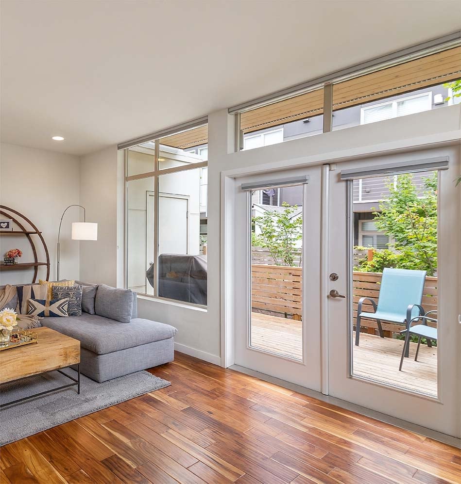 Modern living room with hardwood floors, gray sectional sofa, floor-to-ceiling windows, and sliding glass doors opening to a wooden deck patio