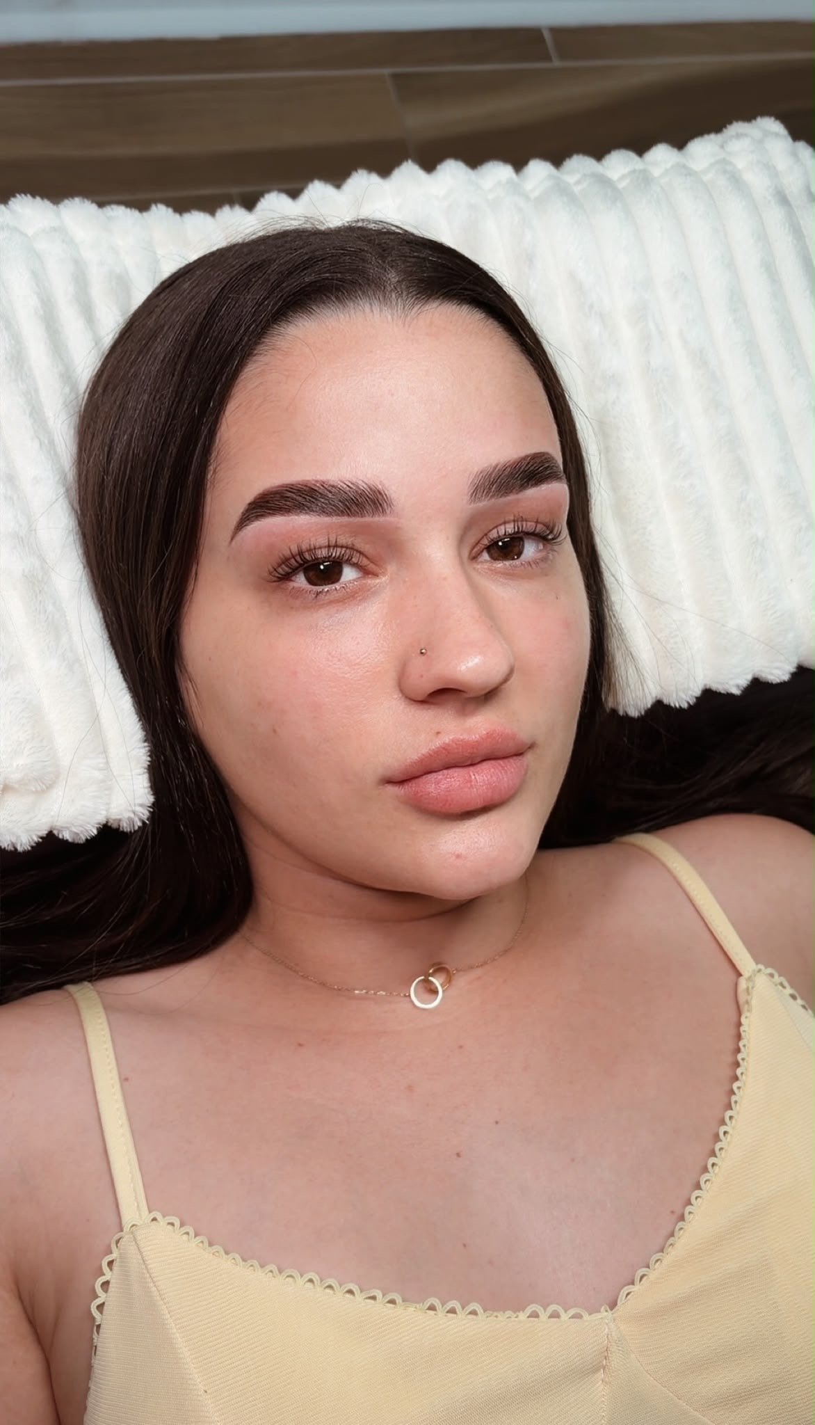 Woman with long dark hair and makeup wearing a pale yellow tank top, resting head on white pillows — private beauty studio experience