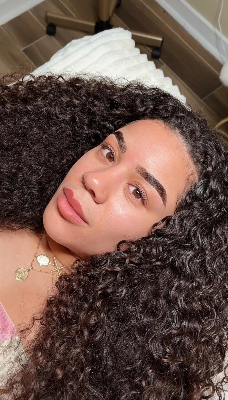 Woman with long curly dark hair wearing a white headwrap, gold jewelry, and natural makeup poses for a selfie indoors