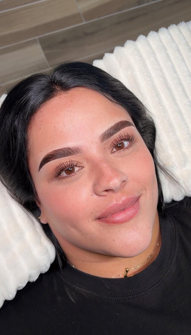 Close-up selfie of a woman with dark hair, defined eyebrows, and pink lipstick, resting her head on a white pillow