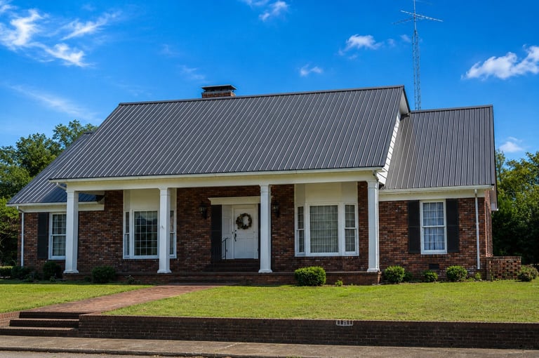 Single-story brick house with metal roof, white trim, covered porch, and manicured lawn