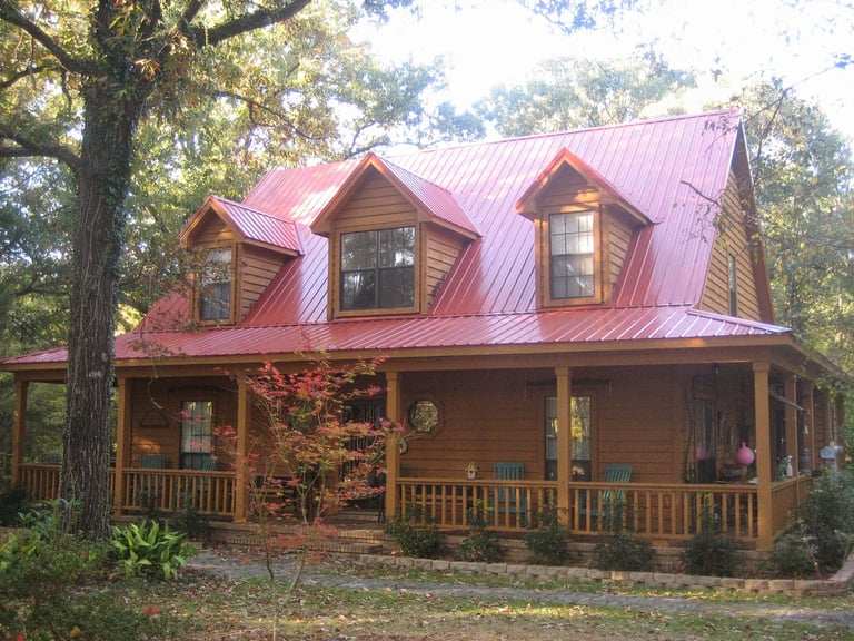 Wooden farmhouse with red metal roof, wraparound porch, and dormer windows surrounded by large trees