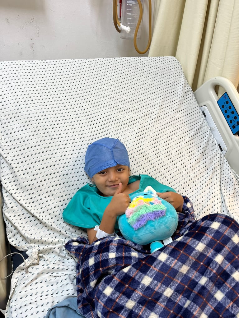 Niña en cama de hospital con gorro quirúrgico azul, sosteniendo un peluche colorido y sonriendo a la cámara