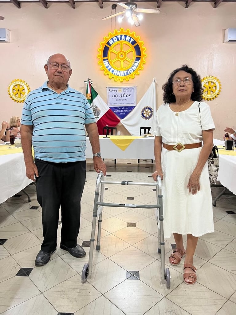 Dos personas mayores de pie en un salón del Club Rotario decorado con los logotipos dorados de Rotary International