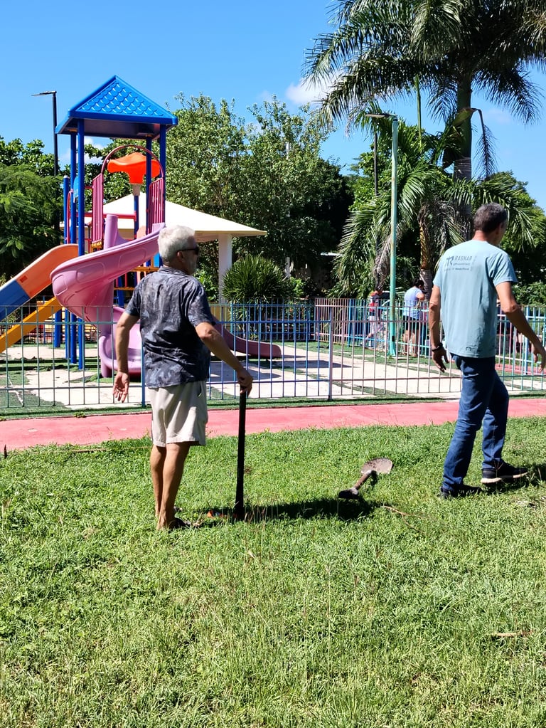 Dos personas jugando cornhole sobre el césped junto a un parque infantil de colores y palmeras durante la jornada