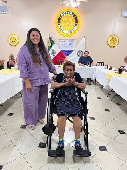 Mujer de blusa morada junto a una señora mayor en silla de ruedas durante una reunión del Club Rotario