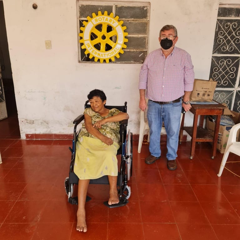 Hombre de playera rosa y cubrebocas junto a una niña en silla de ruedas de blusa amarilla, frente a un muro con un engranaje amarillo