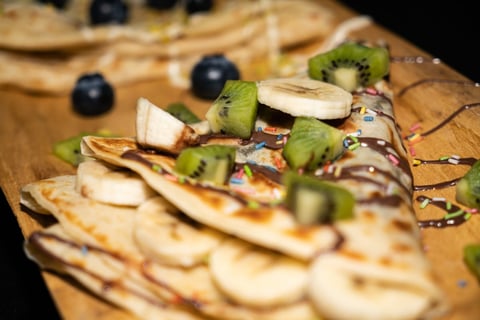 Stack of crepes topped with sliced kiwi, banana, and blueberries on a wooden board