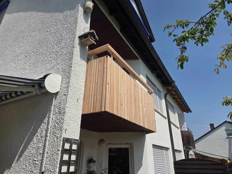 Modern wooden balcony overhang on white-textured residential house exterior with dark roof and foliage visible