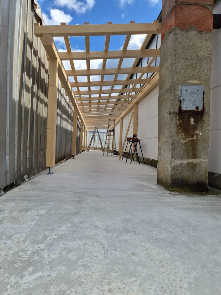 Construction site interior with wooden beam frame structure overhead and concrete floor, featuring ladders and building materials