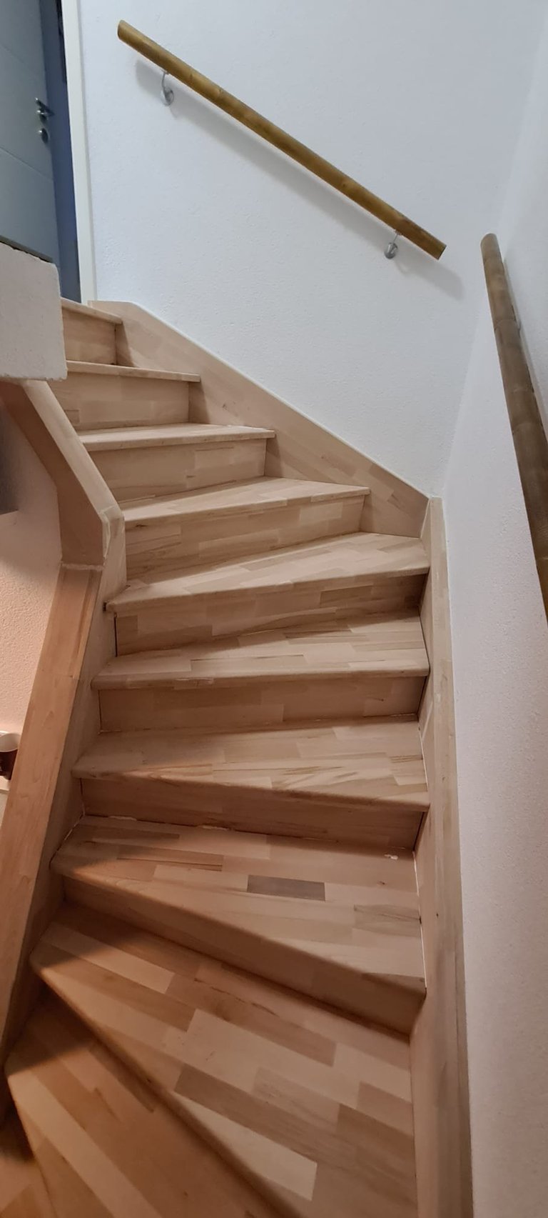 Wooden staircase with light wood steps ascending upward, with a wooden handrail mounted on white wall