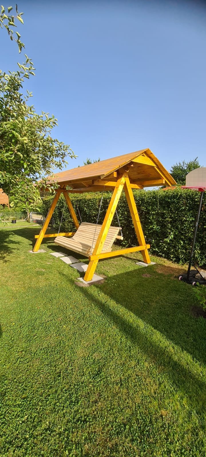 Yellow wooden garden swing with roof canopy in a sunny backyard with green grass and hedge