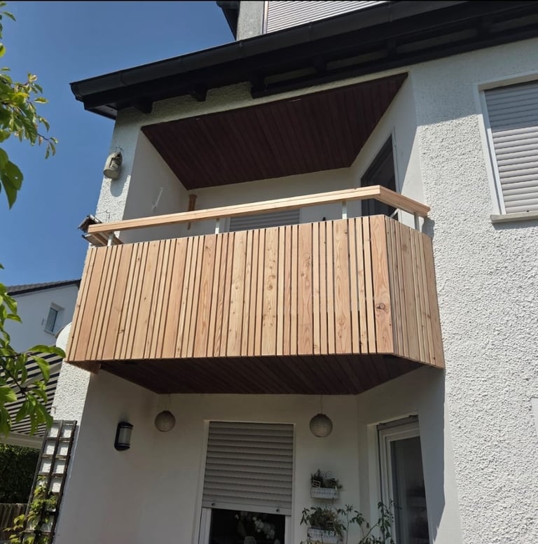Modern white house with wooden balcony featuring metal railings and dark awning, clear blue sky visible above