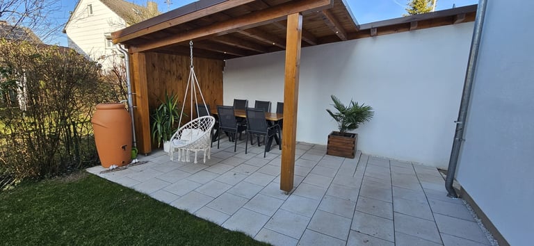 Modern patio with wooden pergola, tiled floor, hanging chair, dining table, potted plants, and white privacy fence