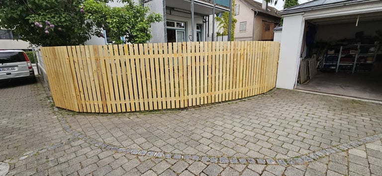 Curved wooden privacy fence installed along a driveway with brick pavement, residential garage visible in background