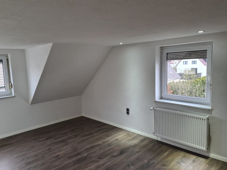 Empty attic room with white sloped ceiling, dark wooden floor, two windows, and wall-mounted radiator