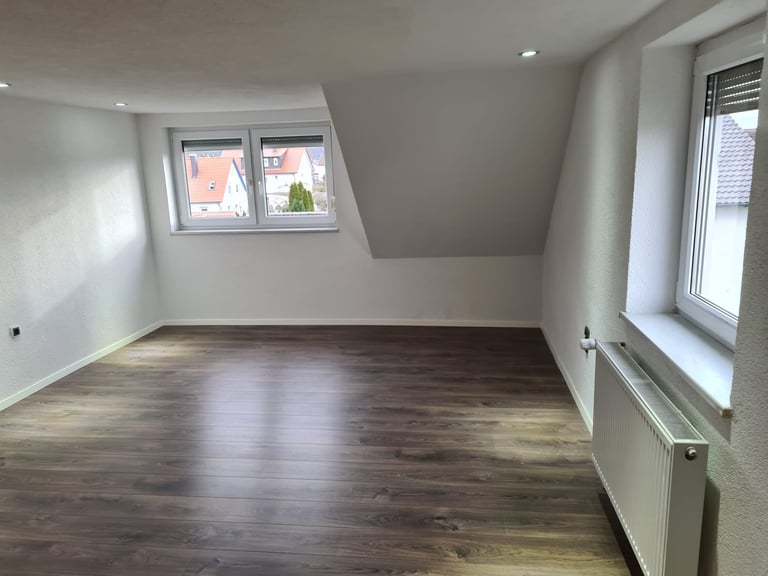 Empty attic room with wooden floor, white walls, sloped ceiling, windows, and radiator
