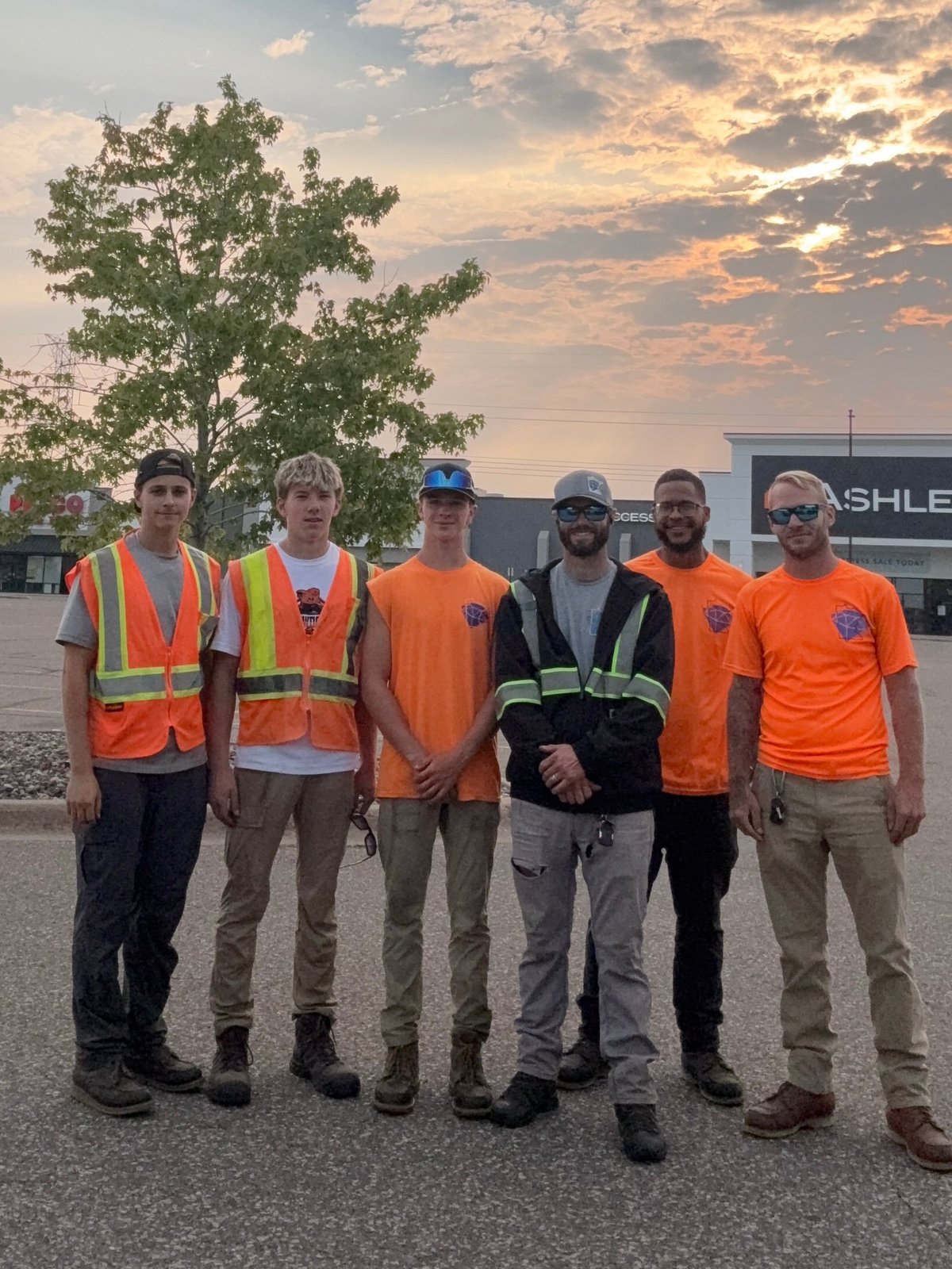 Six workers in safety vests and hard hats standing in a line on an industrial lot at sunset
