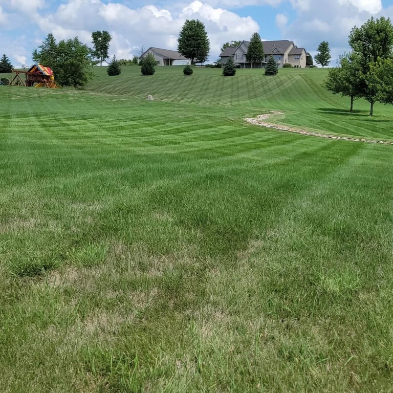 Rolling green hillside with striped mowed lawn patterns, scattered trees, and residential homes in the distance under a partly cloudy sky