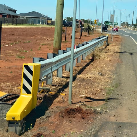 Metal guardrail with yellow safety bumper along rural road construction site with red dirt and suburban buildings visible