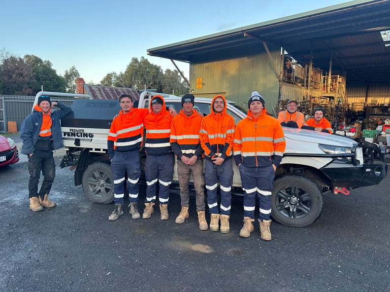 Group of nine workers in orange and navy safety vests standing in front of a white utility truck at an industrial yard