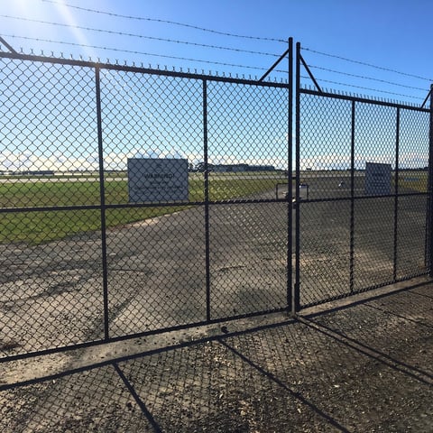 Chain-link fence enclosure with open gate surrounding a dirt and gravel ground area under clear blue sky