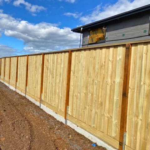 New wooden fence with brown posts along gravel ground beside a building under blue sky with white clouds