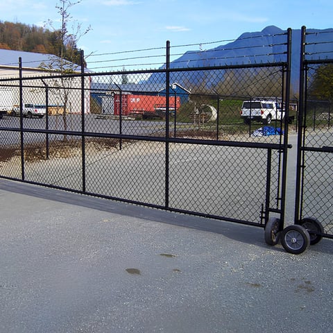 Black chain-link gate with wheels on paved ground, industrial equipment visible beyond fence with mountains in background