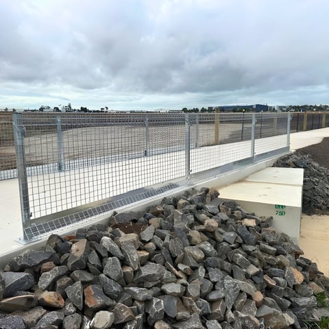 Gray metal mesh safety fencing with rocks piled below on a rooftop overlooking a suburban landscape under cloudy sky