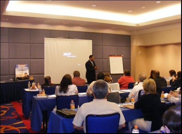 Business presentation in a conference room with attendees seated at blue-draped tables facing a speaker at the front with projection screen