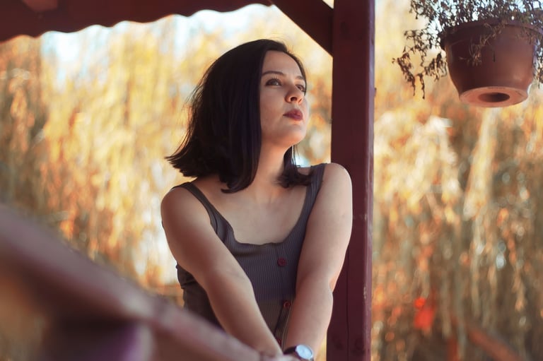 Mujer joven de cabello oscuro con top sin mangas mirando hacia arriba, bajo una estructura rústica de madera con un campo de trigo dorado de fondo, en un momento de introspección