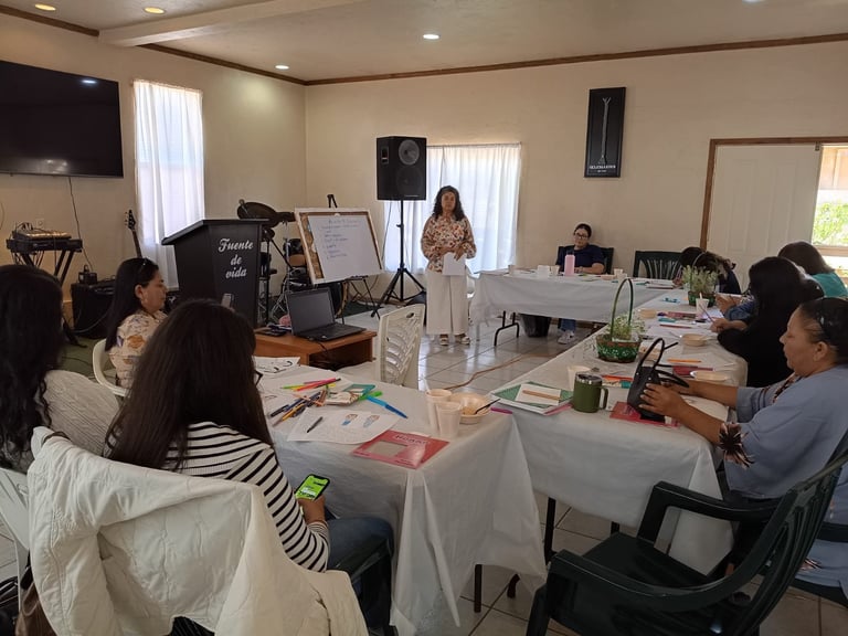Una mujer hablando en un taller en una sala iluminada, con asistentes sentados en mesas y equipo visible al fondo