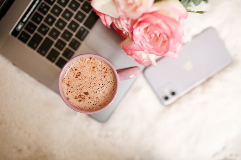 Vista cenital de un espacio de trabajo acogedor con laptop, smartphone, taza rosa con café espumoso y rosas frescas sobre superficie blanca