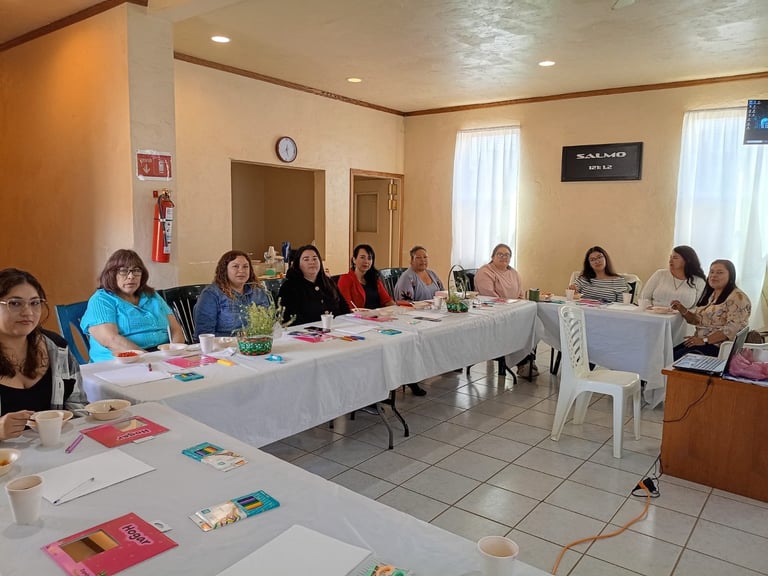 Grupo de mujeres sentadas en mesas largas durante una reunión o taller en una sala con paredes beige, ventanas y un extintor