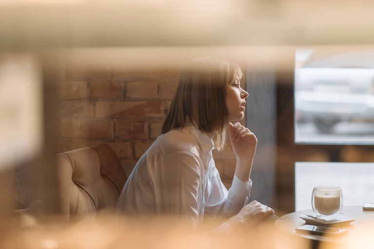 Mujer con camisa blanca sentada junto a una ventana con una taza de café, en un momento de reflexión tranquila y contemplativa