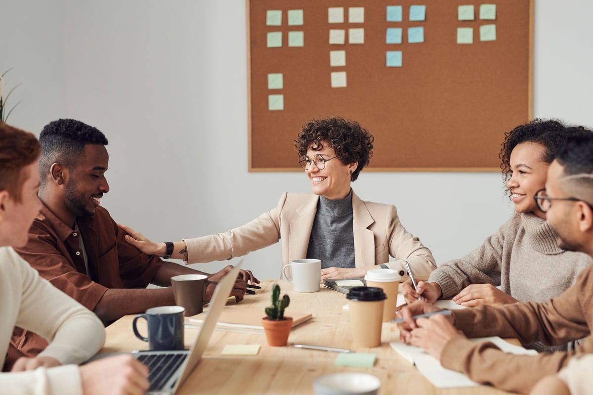 Cinco compañeros de trabajo reunidos alrededor de una mesa de madera en una oficina moderna, sonriendo y conversando en una dinámica de equipo colaborativa