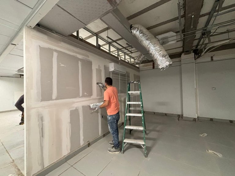 Worker in orange shirt and ladder installing drywall in an industrial space with exposed ceiling beams