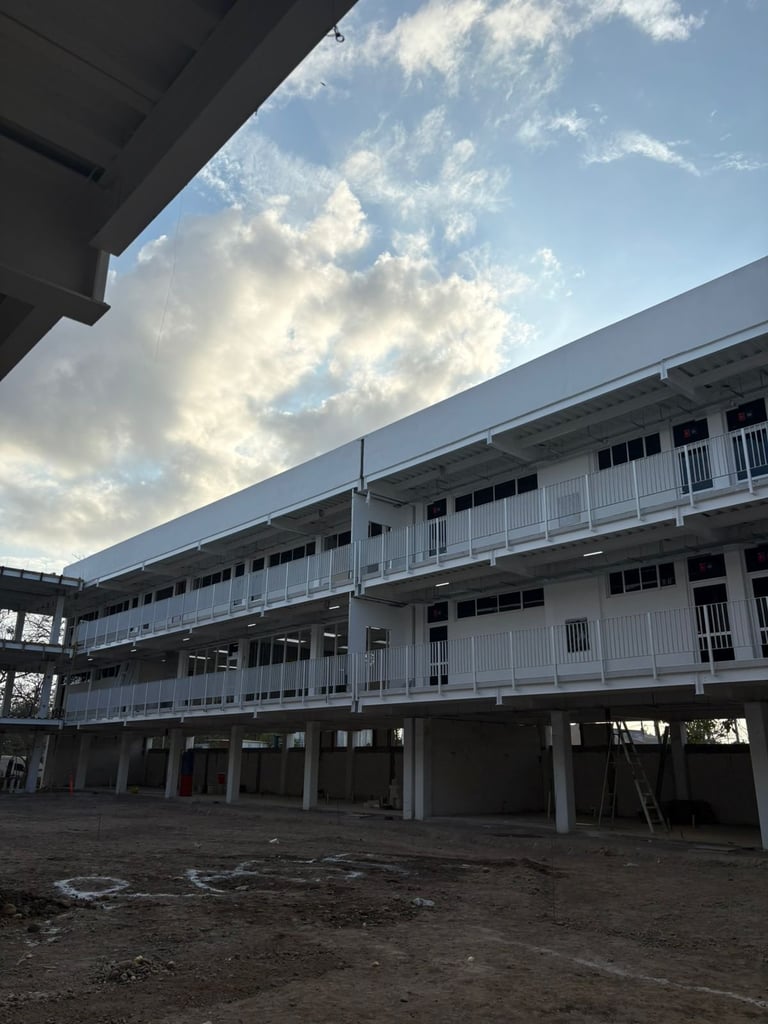 Modern multi-story white building with exposed foundation under construction, viewed from ground level courtyard against cloudy blue sky