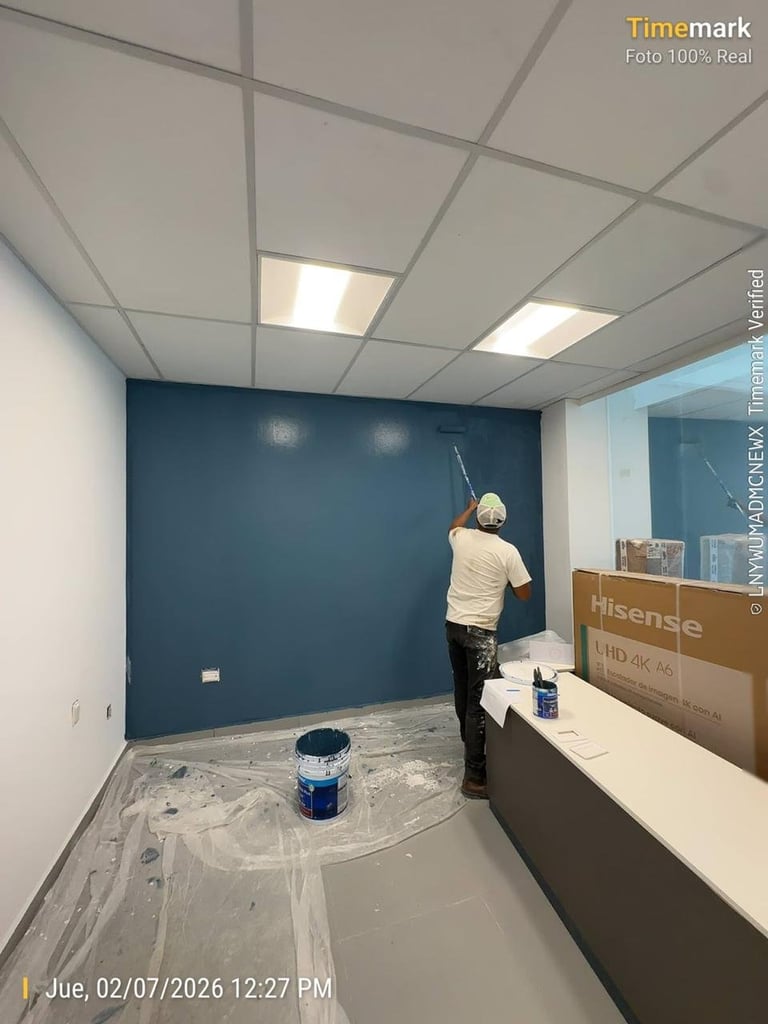 Worker painting a large dark blue wall in an office interior with protective plastic sheeting covering the floor and furniture.